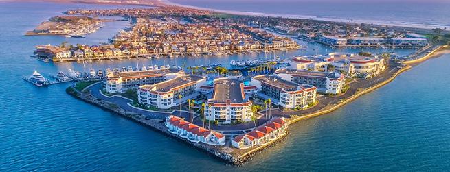 Aerial view of a coastal resort with red-roofed buildings, surrounded by water and boats, under a clear blue sky.
