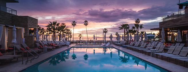 Heated outdoor pool surrounded by lounge chairs under a vibrant sunset sky, with tall palm trees lining the background.